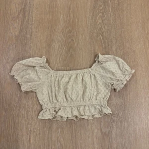 Beige blus med volang från NA-KD - Supersöt beige blus från NA-KD med broderade små hål, puffiga korta ärmar och volangkant nertill. Croppad modell med resår i midjan och ärmslut. Perfekt för dig som gillar romantisk och trendig stil.