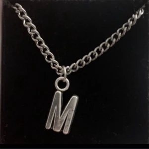 Silverfärgat halsband med olika bokstäver - Stilrent halsband i silverfärg med en kedja och en berlock formad som bokstaven M. Perfekt accessoar för dig som vill bära din initial eller ge bort som personlig present. Kedjan har en klassisk länkdesign och berlocken är enkel men snygg.