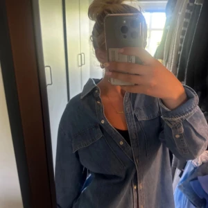 Overshirt i jeans med knappar - Säljer en klassisk blå jeans-overshirt med knappar framtill och två stora bröstfickor. Skjortan har lång ärm och avslappnad passform, perfekt att slänga över en t-shirt. Materialet är robust denim och färgen är mellanblå.