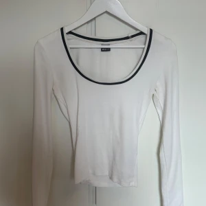 Vit långärmad topp från Gina Tricot - Vit långärmad topp från Gina Tricot med rundad, djup urringning och svart kant runt halsen. Tunn och mjuk i materialet, perfekt för lager-på-lager eller som den är. Enkel och stilren design som funkar till allt.