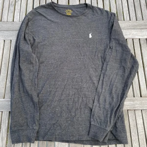 Mörkgrå långärmad tröja Polo Ralph Lauren - Mörkgrå långärmad tröja från Polo Ralph Lauren med klassisk rund hals och broderad vit logga på bröstet. Tillverkad i mjuk bomull som känns skön mot huden. Perfekt för dig som gillar stilren och enkel design.