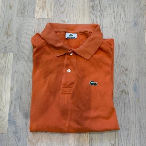 Orange pikétröja från Lacoste - Snygg orange pikétröja från Lacoste med klassisk krage och två knappar framtill. Tröjan har det ikoniska krokodilmärket broderat på bröstet och är tillverkad i bomull som andas. Perfekt för dig som gillar en sportig och stilren look.