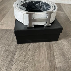 Vitt skärp med silverfärgat H-spänne - Stilrent vitt bälte med präglad struktur och ett stort, silverfärgat H-format spänne. Bältet har en clean look och är tillverkat i skinn. Perfekt accessoar för att lyfta din outfit med en lyxig touch.