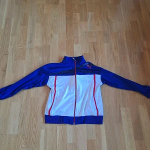 Blå och vit Puma tränings zip tröja - Sportig tränings zip tröja från Puma i blått och vitt med röda detaljer och dragkedja framtill. Tröjan har höga muddar, ståkrage och långa ärmar. Perfekt för dig som gillar en clean och klassisk look med en touch av retrostil. Pris kan såklart diskuteras!