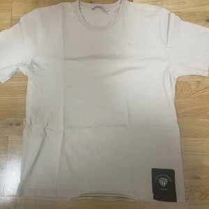 Beige t-shirt från Tiger of Sweden - Snygg beige t-shirt från Tiger of Sweden Jeans med klassisk passform och rund hals. T-shirten har en diskret svart patch med logga nertill på framsidan. Perfekt basic-plagg i mjuk bomull som funkar till allt.