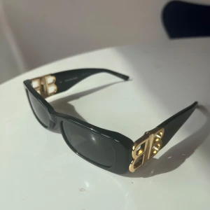 Svarta Balenciaga solglasögon med guldlogo - Snygga svarta solglasögon från Balenciaga med rektangulära bågar och ikoniska guldiga BB-detaljer på sidorna. Glasögonen har mörka linser och en stilren, chunky design 1 BALENCIAGA BB 0096S 001 orginalpris: 2.941,00. Aldrig använt. 