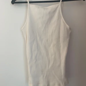 Vit ribbad linnetopp - Säljer en stilren vit ribbad linnetopp med smala axelband. Toppen har en rak passform och är tillverkad i ett mjukt, stretchigt material som sitter skönt mot kroppen. Perfekt att matcha med jeans eller kjol för en clean look.