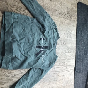 Grön sweatshirt från Calvin Klein Jeans - Snygg grön sweatshirt från Calvin Klein Jeans med stort svart och vitt logotryck på bröstet. Tröjan har rund halsringning och långa ärmar, perfekt för en chill och stilren look. Tillverkad i mjuk bomull som känns skön mot huden.