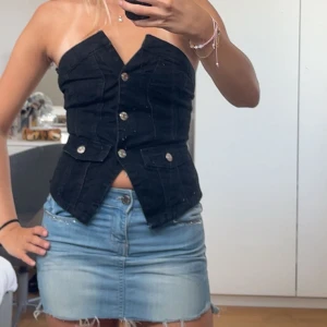 Svart jeans topp med knappar - Säljer en svart, croppad jeans topp med snygg v-ringning och silvriga knappar framtill. Toppen är axelbandslös och har två fickor med lock och knappdetaljer. Perfekt för dig som vill ha en trendig look. Den är aldrig använd💗