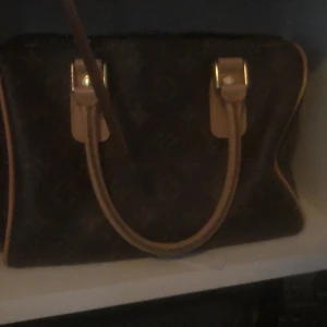  brun monogram handväska - Säljer en klassisk brun handväska med ikoniskt monogrammönster och ljusbruna skinnhandtag. Väskan har guldfärgade metalldetaljer och dragkedja upptill. Perfekt storlek för det viktigaste och snygg att bära i handen.