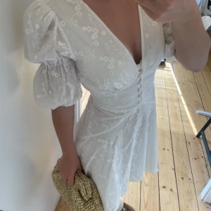 Vit kort klänning med puffärm från adoore - Superfin vit kort klänning med broderade blommor, puffiga korta ärmar och djup v-ringning. Klänningen har knappar framtill och markerad midja som ger en snygg siluett. Perfekt för sommarens alla tillfällen!