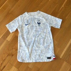 Frankrike Mbappé fotbollströja Nike - Snygg fransk landslagströja från Nike med Mbappé och nummer 10 på ryggen. Tröjan är vit med ljusblått mönster och har korta ärmar. Broderad tupp-logga och Nike swoosh på bröstet. Tillverkad i lätt och ventilerande material, perfekt för match eller träning.