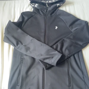 Svart vindjacka från Peace - Svart vindjacka med huva från Peace, dragkedja framtill och två fickor med dragkedja. Snyggt tryck på huvan och liten logga på bröstet. Perfekt för dig som gillar en clean och sportig stil.