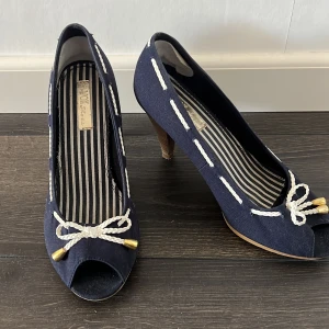 Marinblå pumps med repdetaljer, strl 37 - Snygga marinblå pumps från VOX Shoes med vit repdetalj och guldfärgade ändar framtill. Skorna har öppen tå, randigt innerfoder och hög klack i trälook. Perfekt för dig som gillar nautisk stil och vill sticka ut med coola detaljer.