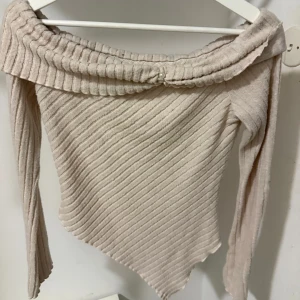 Beige ribbad offshoulder topp - Supersnygg beige ribbad topp med offshoulder och lång ärm. Toppen har en snedskuren nederkant och en söt pärldetalj framtill på kragen. Perfekt för dig som gillar unika detaljer och mjuka material.