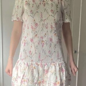 Blommig kortklänning med volang - Supersöt kortklänning i vitt med blommigt mönster i rosa och grönt. Klänningen har korta ärmar, volang nertill och en lätt rynkad struktur i tyget. Perfekt för sommarens alla tillfällen! Osäker på storlek då lappen är bortklippt!!! Men tror L eller XL ( inte i HMs storlekar )