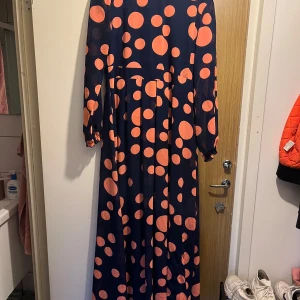Marinblå långklänning med orange prickar - Säljer en maxilång marinblå klänning med stora och små orange prickar. Klänningen har långa ärmar med resår vid handlederna och ett flowy, luftigt fall. Perfekt för dig som gillar färg och mönster! Materialet känns lätt och svalt.