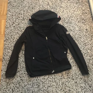 Svart Moncler vindjacka med huva - Svart vindjacka från Moncler med huva och dragkedja framtill. Jackan har Moncler-logga på ärmen och snygga rödvita detaljer på huvan. Perfekt för blåsiga dagar och enkel att matcha med allt. Lätt material och två fickor med knappar.