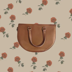  brun handväska i skinn - Snygg brun handväska i halvmåneform med två handtag och stilrena guldfärgade metalldetaljer. Väskan är tillverkad i skinn och har ett rymligt fack med dragkedja upptill. Perfekt för dig som gillar minimalistisk och trendig design.