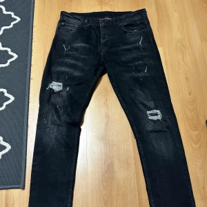 Svarta slitna jeans från Dsquared2 - Svarta jeans från Dsquared2 med coola slitningar och trasiga detaljer på båda benen. Klassisk femficksmodell med smal passform och snygga tvättade effekter. Märkespatch bak i midjan och röda sömmar på insidan. Perfekta för en edgy streetstil.