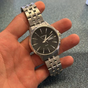 Citizen silverfärgad klocka med svart urtavla - Stilren klocka från Citizen med svart urtavla och dag/datumvisning. Armbandet är i silverfärgat metall och har en klassisk länkdesign. Urtavlan har tydliga indexmarkeringar och smala visare, perfekt för dig som gillar en clean look.