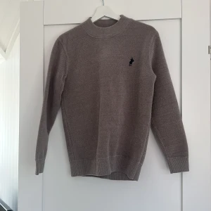 Beige stickad tröja från Ralph Lauren - Säljer en stilren beige stickad tröja från Ralph Lauren med klassisk rund halsringning och broderad logga på bröstet. Tröjan har långa ärmar och ribbade muddar för en snygg passform. Perfekt för dig som gillar en clean och tidlös look.