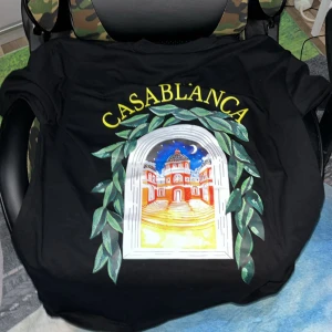 Svart Casablanca t-shirt med tryck - Svart t-shirt från Casablanca med färgglatt tryck av en byggnad och gröna blad, samt gul text på bröstet och ryggen. Klassisk passform och rund hals. Perfekt statement-plagg för dig som gillar unika prints och streetstyle.