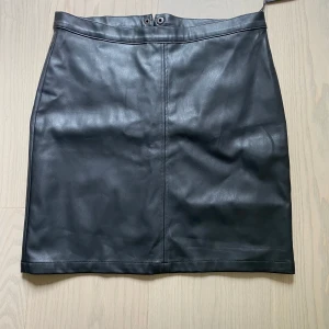 Svart skinnkjol från Black Label S - Snygg svart kort skinnkjol från lager 157 i storlek S. Kjolen har en rak passform, dragkedja och knapp framtill. Perfekt för dig som gillar en stilren och edgy look. Passar till allt från boots till sneakers.