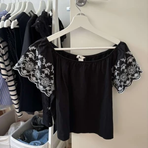 Svart offshoulder blus med broderi - Svart offshoulder blus med vida ärmar och vit broderad spetsdetalj på ärmarna. Blusen har en lös passform och är perfekt för dig som gillar romantiska detaljer och en avslappnad stil.