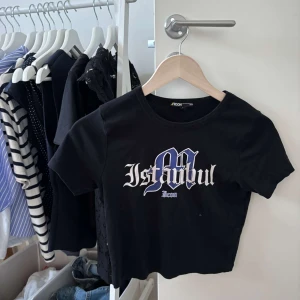 Svart croppad t-shirt Istanbul Icon - Svart croppad t-shirt från Mavi med tryck i vitt och blått där det står 'Istanbul' i gotisk stil. T-shirten har rund halsringning och korta ärmar, perfekt för en cool och avslappnad look.