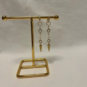 Guldiga örhängen med pärldetalj - Snygga hängande örhängen i guld med små gnistrande stenar, vita pärlor och spetsiga konformade hängen. Örhängena har flera ringar och detaljer som ger en cool och trendig look. Perfekta för dig som vill sticka ut med accessoarer som syns.