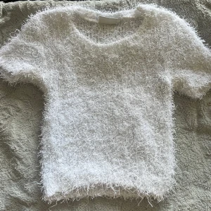 Vit fluffig stickad tröja - Supermjuk och fluffig vit stickad tröja med korta ärmar och rund hals. Tröjan har en cool, lurvig textur som ger en unik look och passar perfekt när du vill sticka ut lite extra. Enkel att styla med både jeans och kjol.