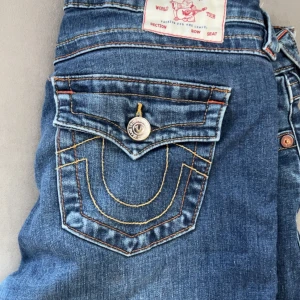 True Religion Joey Low Rise Flare Jeans - Säljer ett par blå True Religion Joey jeans med låg midja och flare-ben. Klassiska kontrastsömmar och stora fickor med snygga detaljer och logoknappar. Perfekta för dig som gillar en retro vibe och vill sticka ut med coola jeans. De har dock en del skada längst ner men det är inget man märker när man har på sig de men utöver de så är de i fint skick. 
