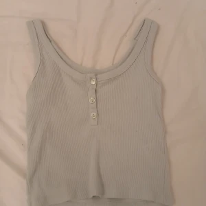 Ljusgrå ribbad linnetopp 157 XS - Säljer en ljusgrå ribbad linnetopp från 157 i storlek XS. Toppen har rundad halsringning och tre dekorativa knappar framtill. Perfekt för en chill och enkel stil, lätt att matcha med jeans eller shorts.