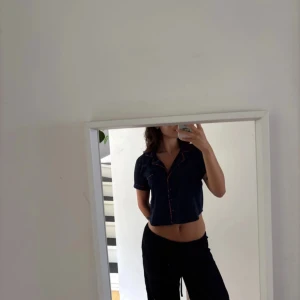 Mörkblå croppad skjorta med text - Säljer en mörkblå croppad skjorta med kort ärm och röda detaljer längs krage och ärmslut. På ryggen står det 'No Peeking' i röd text. Skjortan har knappar framtill och en avslappnad passform. Perfekt för dig som vill sticka ut lite extra.