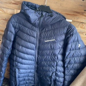 Mörkblå dunjacka Peak Performance M - Snygg mörkblå dunjacka från Peak Performance med huva och dragkedja framtill. Jackan har quiltad design och logga på bröstet. Perfekt för kalla dagar och enkel att matcha med det mesta. Klassisk look och riktigt skön passform.har några små hål som är lätta att laga och täcka (syns troligen inte)