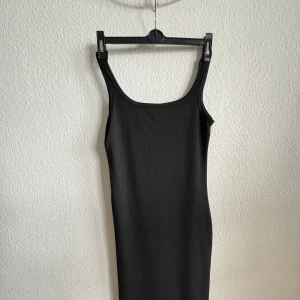 Svart ribbad ärmlös klänning - En stilren svart ribbad klänning med tight passform och ärmlös design. Klänningen har breda axelband och en rundad halsringning. Perfekt för dig som gillar minimalistisk och clean stil. Materialet är stretchigt och följsamt.