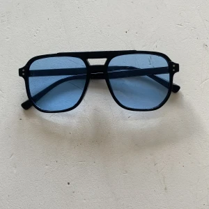 Svarta pilotbågar med blåtonade glas - Snygga solglasögon med svart pilotformad båge och blåtonade glas. Ramen är i plast och har en cool retrovibe med dubbla nitar på sidorna. Perfekt accessoar för dig som vill sticka ut med en klassisk men trendig look.
