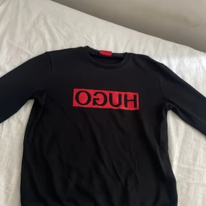 Svart tröja med röd HUGO-logga - Svart långärmad tröja från Hugo Boss med stor röd HUGO-logga framtill. Tröjan har rund halsringning och är i en klassisk, normal passform. Perfekt för dig som gillar stilrena och snygga statement-plagg.