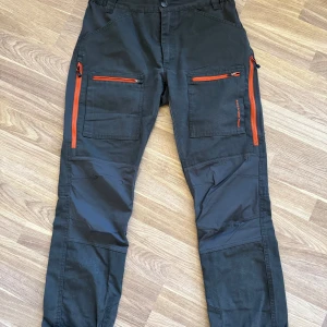 Fritidsbyxor från High Mountain - Mörkgröna fritidsbyxor från High Mountain med orangea detaljer. Flera fickor. Storlek 40.