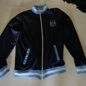 Manchester City retro track jacket XL - Snygg retro track jacket från Manchester City i mörkblått med ljusblå och vita detaljer. Jackan har klubbens klassiska emblem på bröstet, hel dragkedja och ribbade muddar med ränder. Den är i mycket bra skick! Obs! Står xl men passar M-XL