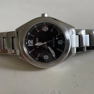 Citizen Eco-Drive klocka i silver - Citizen Eco-Drive armbandsur med svart urtavla och datumvisning. Klockan har en robust länkad boett och armband i silverfärgat rostfritt stål. Snygg och stilren design med tydliga siffror och markeringar. Perfekt för dig som gillar klassiska accessoarer.