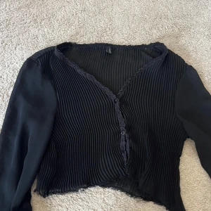 Svart ribbad blus i mesh - Säljer en svart blus från FREE/QU med ribbad, transparent och släta långärmade ärmar. Toppen har v-ringning med spetskant, knappar framtill och en lätt croppad, vågig nederkant. Perfekt för dig som gillar unika detaljer och stilren look.