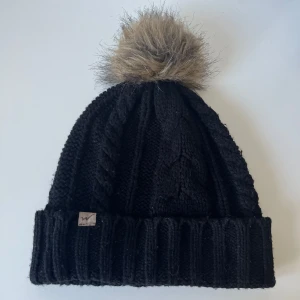 Svart stickad mössa med fuskpälsboll - Svart stickad mössa med grov kabelstickning och bred uppvikt kant. På toppen sitter en stor, fluffig fuskpälsboll i beige/bruna toner.