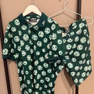 Grön mönstrad set från J.Lindeberg - Snygg grön pikétröja från J.Lindeberg med vit logomönster över hela tröjan. Klassisk krage med broderad logga på kragspetsen och knäppning framtill. Skön passform och perfekt för dig som gillar sportig stil. Matchande shorts.Golf kläder 