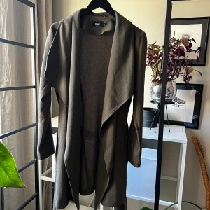 Olivgrön kappa från ONLY med bälte - Snygg olivgrön kappa från ONLY med avslappnad passform och bälte i midjan. Jackan har långa ärmar, öppen front och mjukt, strukturerat tyg. Perfekt för lager-på-lager och en clean look till höst och vår.