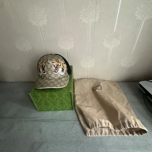 Gucci keps  tiger - Snygg keps från Gucci med klassiskt GG-mönster i beige och brunt, broderade tigrar framtill och svart mesh baktill. Justerbar rem med grönt och rött band. Perfekt statement-piece för dig som vill sticka ut.