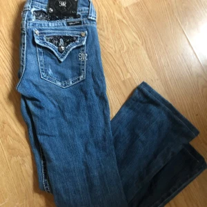 Miss Me blå bootcut jeans med detaljer - Snygga blå jeans från Miss Me med bootcut-modell. Jeansen har dekorativa fickor med broderier, nitar och glittriga detaljer samt en läderpatch bak. Tydliga kontrastsömmar och coola metallknappar ger extra edge. Perfekta för dig som gillar statement-jeans.
