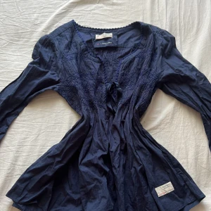 Mörkblå blus med knyt från Odd Molly - Supersöt mörkblå blus från Odd Molly med broderade detaljer och knyt framtill. Blusen har lång ärm, v-ringning och smockad överdel som ger en snygg passform. Perfekt för dig som gillar boho-vibbar och unika detaljer.