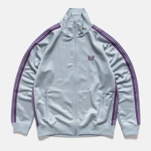 Needles Japan Track Jacket - Snygg ljusblå track jacket från Needles Japan med lila ränder längs ärmarna och broderad fjäril på bröstet. Jackan har dragkedja framtill, höga muddar och två fickor med dragkedja. Perfekt för en sportig och avslappnad stil. Cond 9,5/10